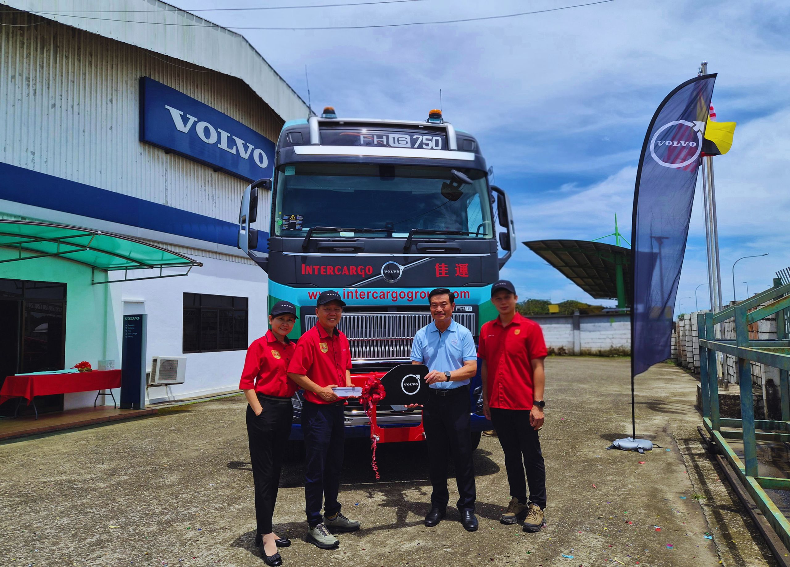 Five people in red uniforms pose with a new Volvo truck during a ribbon-cutting ceremony at a Volvo dealership.